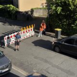 Ein schwarzes Auto steht vor einer Absperrung, an der mehrere Ordner teils in orangefarbenen Warnwesten stehen