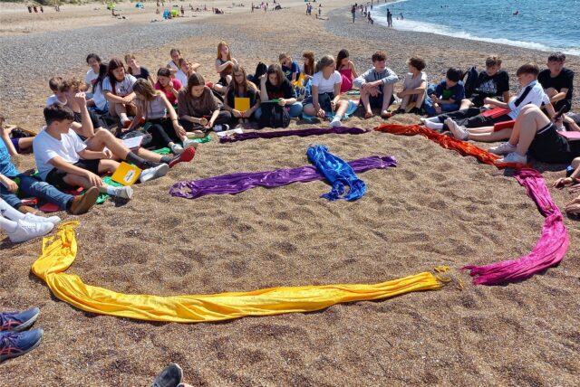 Die Konfirmanden der evangelischen Kirche Kamen 2024 sitzen im Kreis an einem Strand. In der Mitte haben sie bunte Tücher auf den Boden gelegt, die einen Kreis mit einem Kreuz in der Mitte bilden.