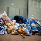 Ein Obdachloser schläft in einer Fußgängerzone eingepackt in Decken und zwischen Plastiktüten auf dem Boden.