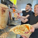 Drei Männer arbeiten hinter der Theke eines Döner-Restaurants in Unna. Einer hält lächelnd eine gefüllte Teigtasche nach vorn.