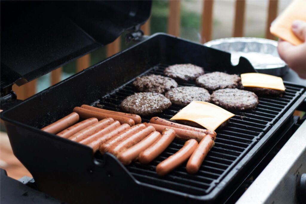 Auf einem Grillrost werden zahlreiche Würstchen, fünf Burgerpattys und zwei Scheiben Käse gegrillt