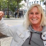 Anja Schwerdtfeger, eine Frau mit blonden Haaren, lehnt sich an einen Baum und lächelt in die Kamera. Sie trägt eine weiße Jeansjacke und einen grauen Pullover.