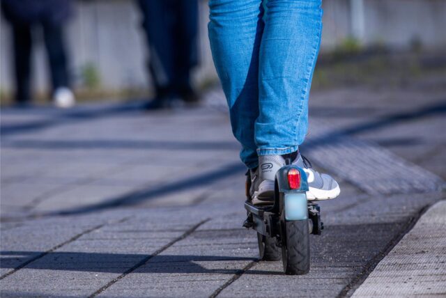 Ein E-Scooter ist von hinten zu sehen. Darauf stehen zwei Beine mit blauer Jeans und Turnschuhen an. Die Füße stehen hintereinander. Der Hintergrund ist verschwommen. Der E-Scooter steht auf einem Weg mit grauen Steinplatten.