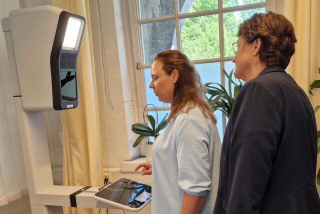 Zwei Frauen stehen vor einem Gerät mit Bildschirm in einem Raum mit Fenstern und Pflanzen. Die Frau im hellblauen Oberteil tippt auf den Bildschirm des Geräts.