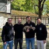 (v.l.) Philipp Steinke, Torsten Weichert, Phil Couwenbergs mit Hund Anton auf dem Arm und Deniz Özdemir stehen auf dem Gelände der Galopprennbahn in Dortmund Wambel. Hinter ihnen befindet sich der Außengastronomiebereich mit verschlossenen Rollos.