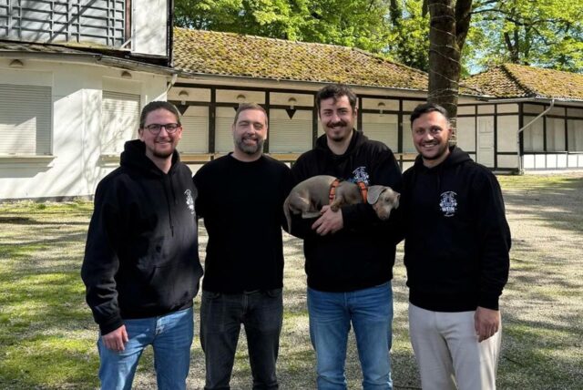 (v.l.) Philipp Steinke, Torsten Weichert, Phil Couwenbergs mit Hund Anton auf dem Arm und Deniz Özdemir stehen auf dem Gelände der Galopprennbahn in Dortmund Wambel. Hinter ihnen befindet sich der Außengastronomiebereich mit verschlossenen Rollos.