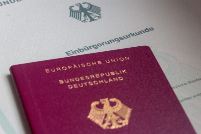 Im Vordergrund ist der obere Teil eines roten Reisepasses zu sehen. Darauf steht in goldenen Buchstaben Europäische Union, darunter in kleinerer Schrift Bundesrepublik Deutschland. Darunter ist der Bundesadler abgedruckt. Der Pass liegt auf einem Dokument, auf dem Einbürgerungsurkunde steht.