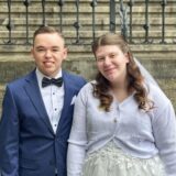Ein junges Paar posiert in Hochzeitskleidung vor einer Steintreppe, die mit Blumen geschmückt ist. Der Mann trägt einen dunkelblauen Anzug mit Fliege, während die Frau ein weißes Kleid mit einem passenden Cardigan und Schleier trägt.