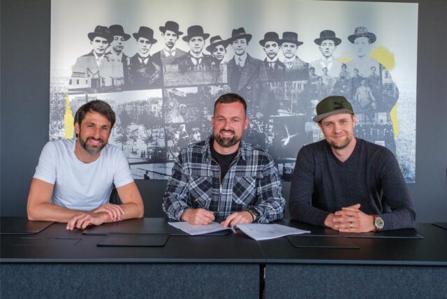 Thomas Broich, Felix Hirschnagl und Paul Schaffran sitzen an einem Schreibtisch und gucken geradeaus in die Kamera.