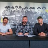 Thomas Broich, Felix Hirschnagl und Paul Schaffran sitzen an einem Schreibtisch und gucken geradeaus in die Kamera.
