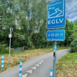 Das Bild zeigt einen Abschnitt des Emscherwegs, einem Fußgänger- und Fahrradweg, gekennzeichnet durch blaue Schilder der Emschergenossenschaft EGLV. Ein Schild weist darauf hin, dass auf dem Weg kein Winterdienst erfolgt.