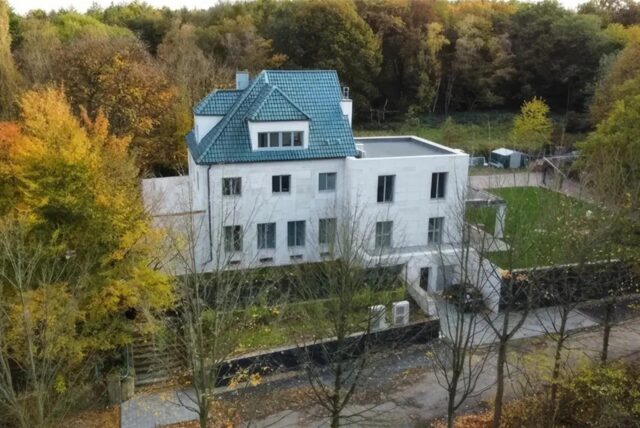 Ein mehrstöckiges, weißes Gebäude mit blauem Dach ist von Bäumen umgeben. Im Hintergrund befindet sich ein Wald mit herbstlichem Laub.