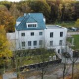 Ein mehrstöckiges, weißes Gebäude mit blauem Dach ist von Bäumen umgeben. Im Hintergrund befindet sich ein Wald mit herbstlichem Laub.