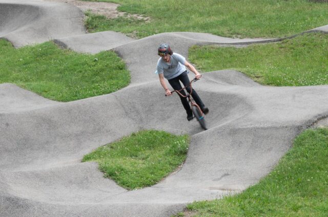 Ein Radfahrer ist auf einer asphaltierten BMX-Bahn zu sehen. Die Bahn ist durch Hügel und Kurven gekennzeichnet.