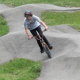 Ein Radfahrer ist auf einer asphaltierten BMX-Bahn zu sehen. Die Bahn ist durch Hügel und Kurven gekennzeichnet.