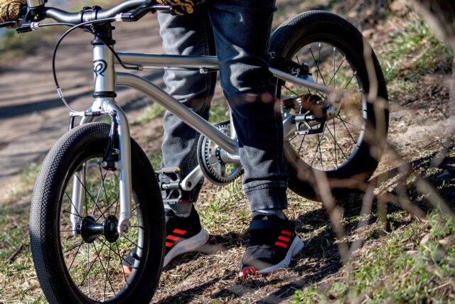 Ein Junge mit schwarz-orangefarbenen Turnschuhen sitzt auf einem silberfarbenen, kleinen Fahrrad.