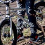 Ein Junge mit schwarz-orangefarbenen Turnschuhen sitzt auf einem silberfarbenen, kleinen Fahrrad.