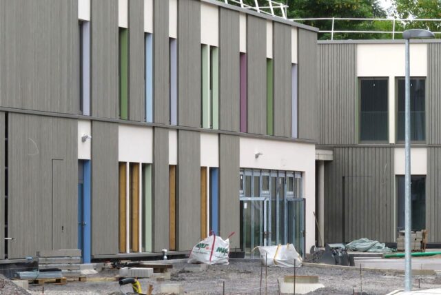 Die Außenfassade der neuen Schule am Hertinger Tor ist geprägt von einer grauen Holzlattung, weißen Putzflächen und bunt gestrichenen Fensterleibungen.