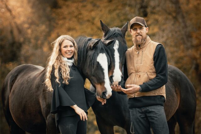 Auf dem Bild sind eine Frau und ein Mann neben vier Pferden auf einem Reitplatz im Freien zu sehen. Die Personen lächeln in die Kamera, während die Pferde unterschiedlich schauen.