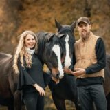 Auf dem Bild sind eine Frau und ein Mann neben vier Pferden auf einem Reitplatz im Freien zu sehen. Die Personen lächeln in die Kamera, während die Pferde unterschiedlich schauen.