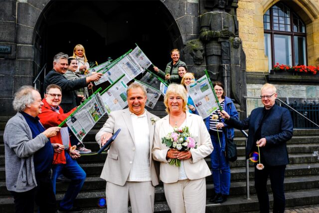 Ralf Wiethaup und Doris Hübecker stehen nach ihrer Trauung vor dem Rathaus in Recklinghausen, im Hintergrund gratulieren die Kollegen der Recklinghäuser Zeitung mit Zeitungsstöcken und Blumen