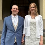 Ein frisch vermähltes Paar posiert Händchen haltend vor einem Gebäude. Die Frau trägt ein weißes Kleid mit Spitze und eine kurze weiße Jacke, während der Mann einen blauen Anzug mit beiger Weste trägt.