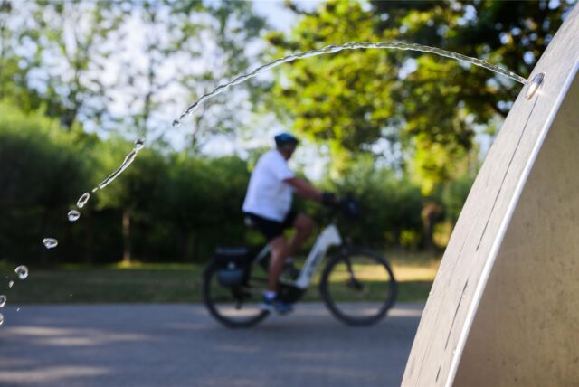 Im Vordergrund ist ein Wasserhahn zu sehen, aus dem Wasser austritt. Im Hintergrund fährt eine Person mit einem E-Bike auf einem asphaltierten Weg entlang.