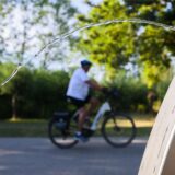 Im Vordergrund ist ein Wasserhahn zu sehen, aus dem Wasser austritt. Im Hintergrund fährt eine Person mit einem E-Bike auf einem asphaltierten Weg entlang.