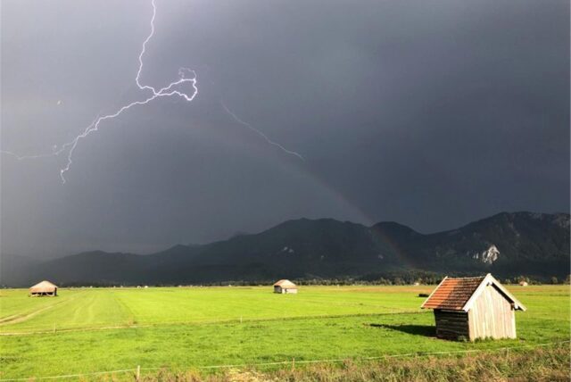 Ein Blitz ist am Himmel über den Bergen zu sehen, im Vordergrund eine grüne Wiese.