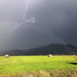 Ein Blitz ist am Himmel über den Bergen zu sehen, im Vordergrund eine grüne Wiese.
