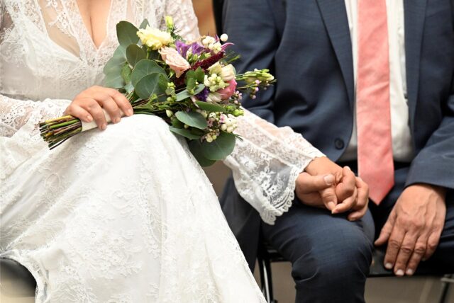 Ein Ehepaar sitzt nebeneinander. Die Braut hält einen Blumenstrauß in der Hand. Ihre Gesichter sind nicht zu sehen.