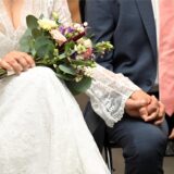 Ein Ehepaar sitzt nebeneinander. Die Braut hält einen Blumenstrauß in der Hand. Ihre Gesichter sind nicht zu sehen.
