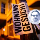 Ein Zettel mit der Aufschrift „Wohnung gesucht“ hängt vor Wohnhäusern.