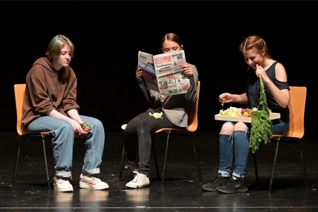 Drei junge Frauen sitzen auf Stühlen vor einem dunklen Hintergrund. Eine liest Zeitung, eine hält Gemüse in der Hand und die dritte isst eine Karotte. Drei junge Frauen sitzen auf Stühlen vor einem dunklen Hintergrund. Eine liest Zeitung, eine hält Gemüse in der Hand und die dritte isst eine Karotte.