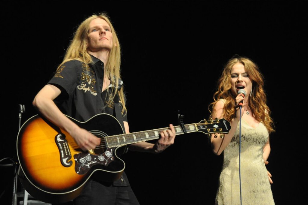 Auf dem Bild sind ein Gitarrist mit langen blonden Haaren und eine Sängerin mit lockigen braunen Haaren auf einer Bühne zu sehen. Die Sängerin singt in ein Mikrofon, während der Gitarrist eine Akustikgitarre spielt. Auf dem Bild sind ein Gitarrist mit langen blonden Haaren und eine Sängerin mit lockigen braunen Haaren auf einer Bühne zu sehen. Die Sängerin singt in ein Mikrofon, während der Gitarrist eine Akustikgitarre spielt.