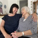 Zwei Frauen sitzen auf einem Sofa und lachen sich an. Eine der Frauen hat dunkles Haar, die andere graues.