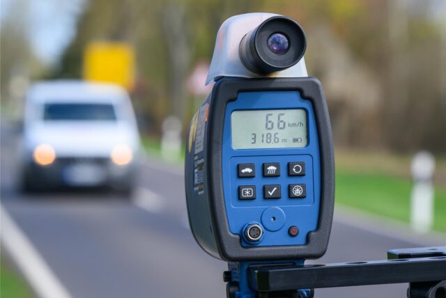 Eine mobile Geschwindigkeitsmessanlage zeigt eine gemessene Geschwindigkeit von 66 km/h an. Im Hintergrund ist ein unscharfes Auto auf einer Landstraße zu sehen.