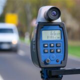 Eine mobile Geschwindigkeitsmessanlage zeigt eine gemessene Geschwindigkeit von 66 km/h an. Im Hintergrund ist ein unscharfes Auto auf einer Landstraße zu sehen.