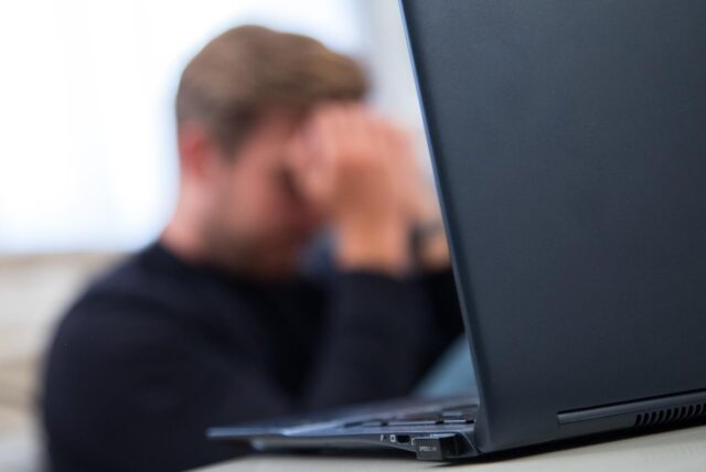 Ein Mann sitzt mit den Händen vor dem Gesicht verzweifelt hinter einem Laptop.