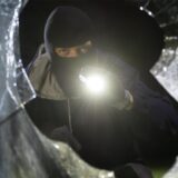 Ein maskierter Mann hält eine Taschenlampe in der Hand und steht vor einer zerschlagenen Scheibe.