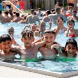 Einige Kinder posieren in einem Schwimmbecken in einem Freibad für ein Foto und lachen in die Kamera. Im Hintergrund befinden sich viele weitere Personen im Wasser.