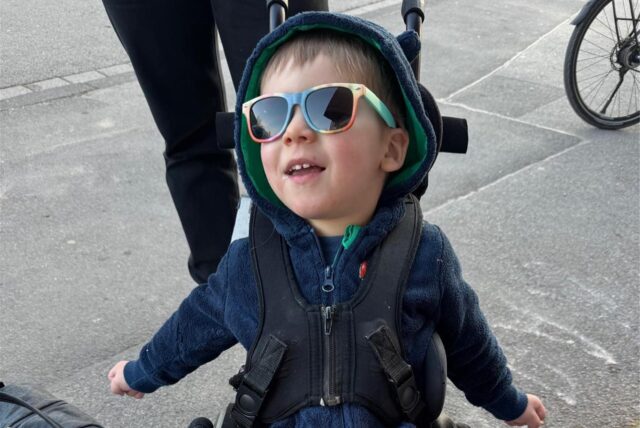 Ein kleiner Junge sitzt im Rollstuhl. Er trägt eine Sonnenbrille.