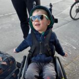 Ein kleiner Junge sitzt im Rollstuhl. Er trägt eine Sonnenbrille.