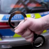 Eine Hand hält eine silberne Handschelle vor dem Heck eines Streifenwagens der Polizei. Die Rücklichter und die Aufschrift „POLIZEI“ auf dem Fahrzeug sind erkennbar.