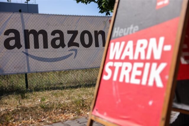 Ein Amazon-Logo ist auf einem Zaun im Hintergrund zu sehen. Im Vordergrund steht ein Schild mit der Aufschrift „Heute Warnstreik“.