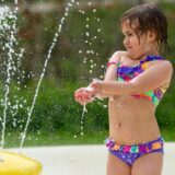 Kleines Mädchen in bunter Badebekleidung spielt mit einer Wasserfontäne im Freien.