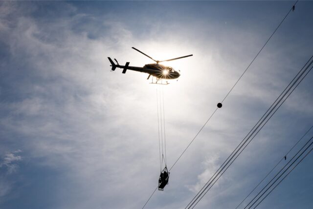Ein Hubschrauber transportiert eine Gondel unterhalb von Stromleitungen. Die Aufnahme wurde gegen die Sonne gemacht, wodurch eine Silhouette entsteht.