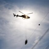Ein Hubschrauber transportiert eine Gondel unterhalb von Stromleitungen. Die Aufnahme wurde gegen die Sonne gemacht, wodurch eine Silhouette entsteht.