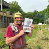 Björn Volland steht mit seinem Comic vor der Holz-Erde-Mauer im Römerpark Bergkamen.