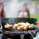 Grill mit bratenden Würstchen im Vordergrund, unscharfe Menschen im Hintergrund auf Picknickdecke